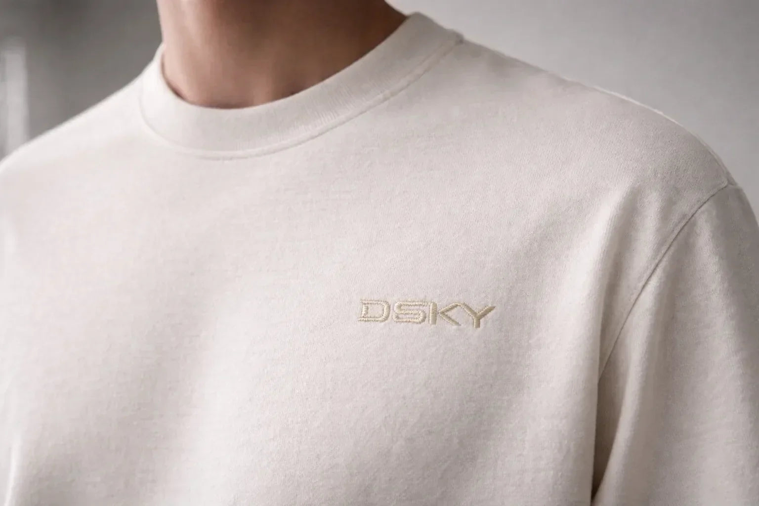 Close-up van een model dat een off-white DSKY T-shirt draagt met subtiele tonale borduring op de borst, minimalistische premium streetwear.