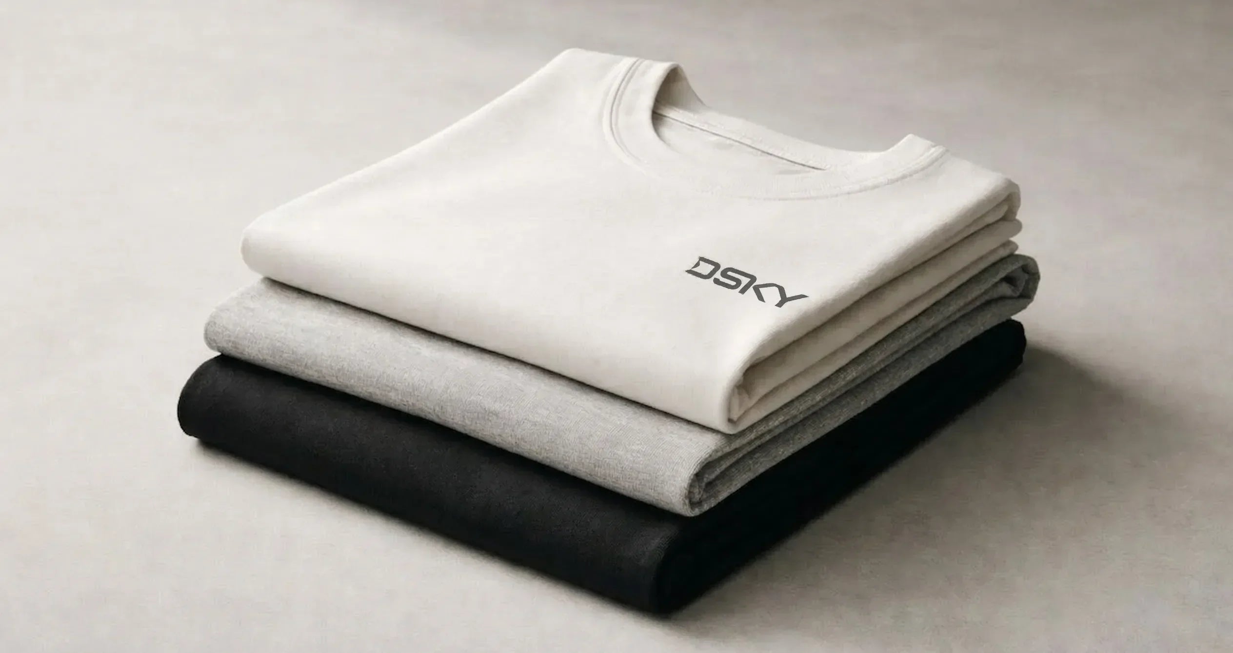 Gestapelde premium katoenen T-shirts in neutrale tinten met subtiel DSKY logo, minimalistische studiofotografie gericht op materiaal en stofstructuur.