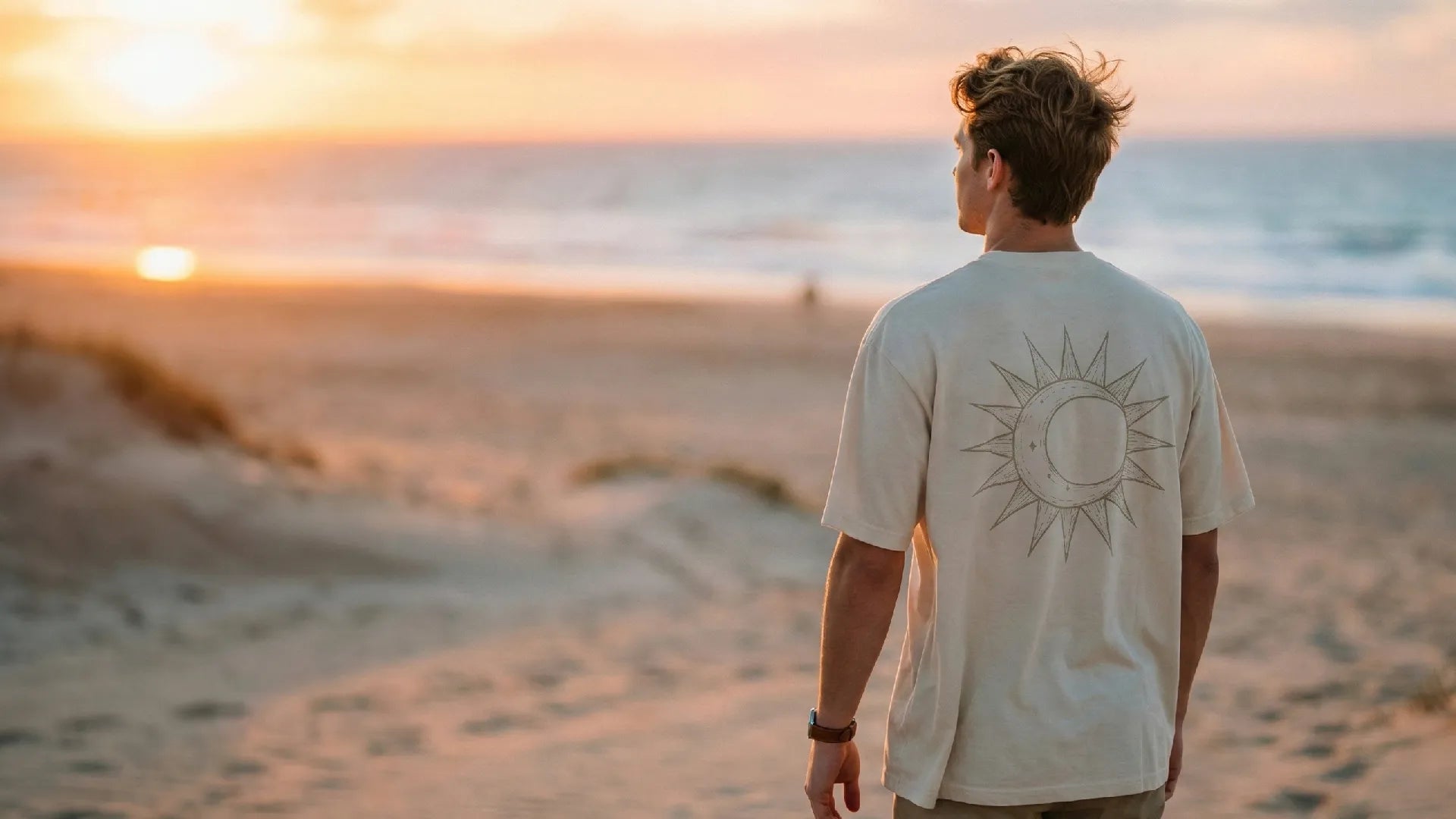 Man in a DSKY Clothing beige t-shirt standing on a sandy beach at sunset, looking towards the ocean.